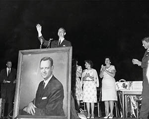Black and white photo from the Broyhill exhibit of a man standing on stage behind a large portrait