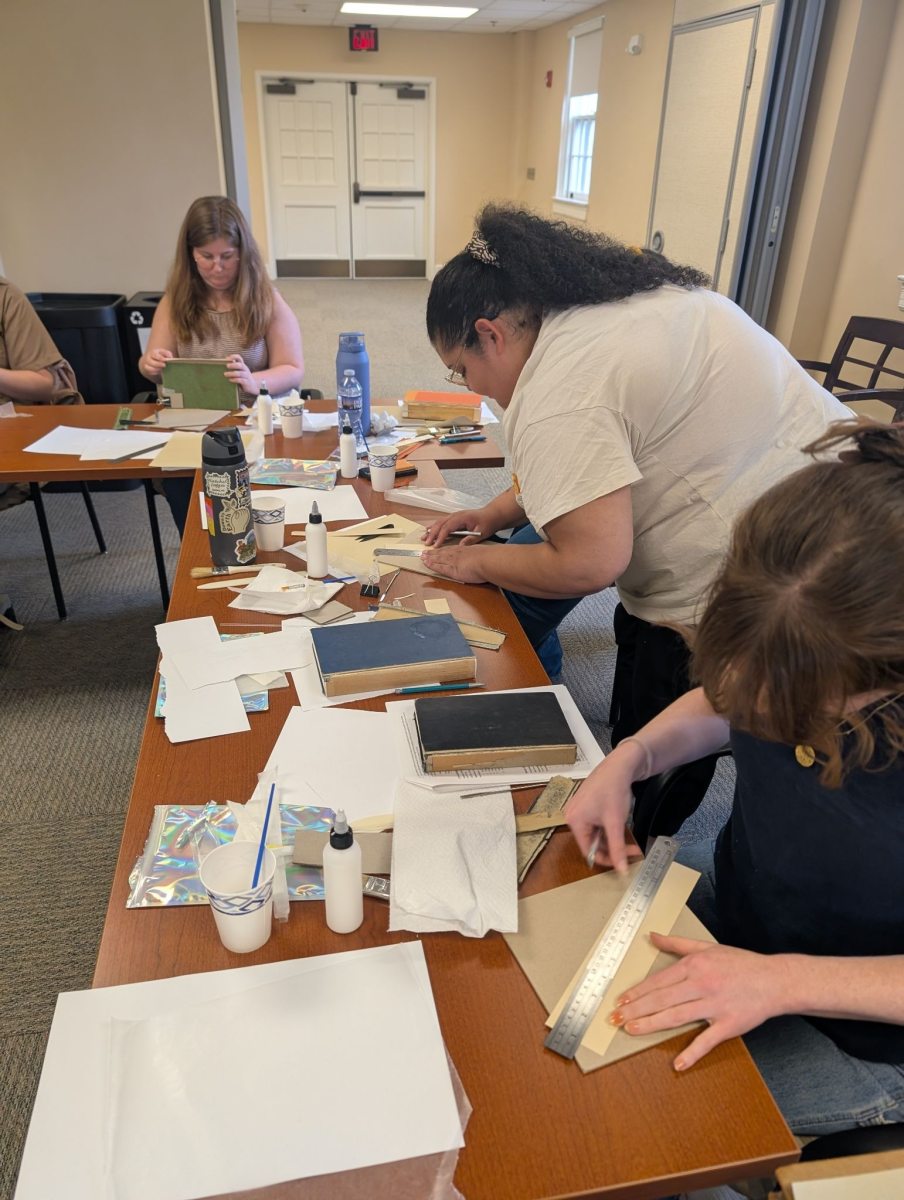 Hannah Helmey and Celine Shay working at a long table that is covered with books and book repair supplies, such as glue, papers, and brushes. They are using rulers to make measurements in preparation for spine replacement.