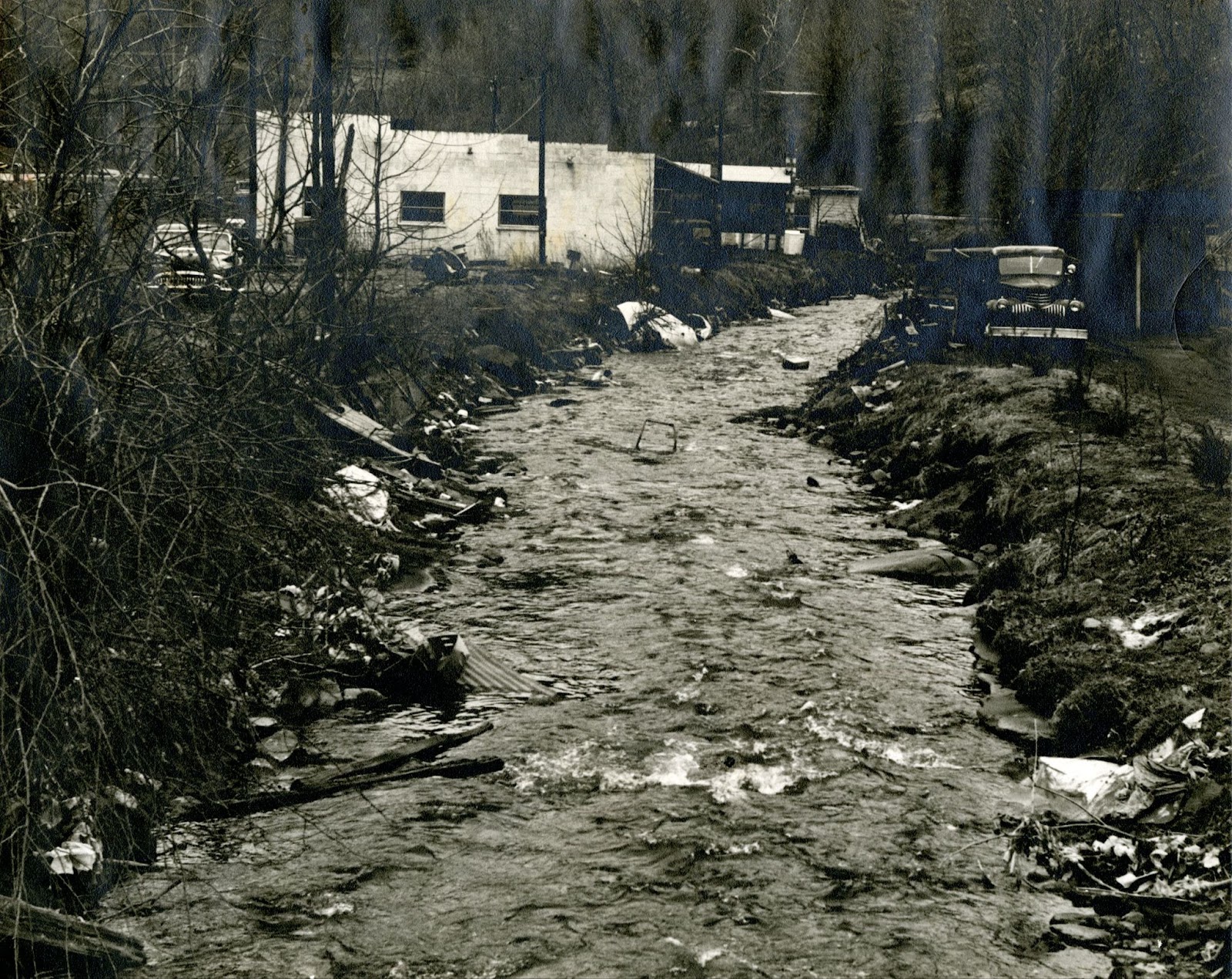Black and white photo of creek running behind a building. Trash and debris is visible on the banks.