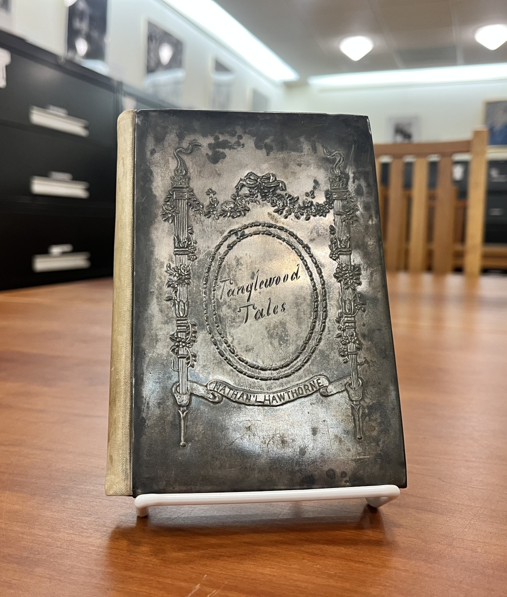 An antique book titled "Tanglewood Tales" by Nathaniel Hawthorne. The front cover features a tarnished, metallic silver plate embossed with neoclassical pillars, floral garlands, and an oval border surrounding the cursive title "Tanglewood Tales." A banner below reads "Nathan'l Hawthorne"