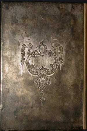 Close-up of the back cover plate of an antique book. The metallic, heavily aged silver surface features a single, centered decorative motif. The embossed design includes an elegant neoclassical crest made of scrolling acanthus leaves, flaming torches with flowing ribbons, and a detailed circular wreath dangling from the bottom center